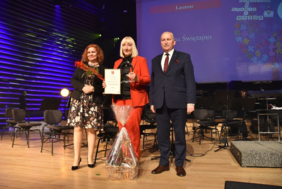 Pokaż zdjęcie: Scena filharmonii Olsztyńskiej. Od lewej stoją ruda kobieta w sukience w kwiaty i marynarce, po środku: platynowa blondynka w czerwonym garniturze i czarnej bluzce, z dyplomem i statuetką w dłoniach oraz koszem słodkości u stóp. Po prawej ubrany w