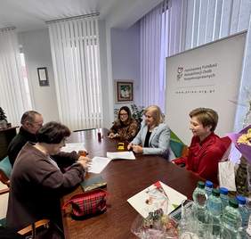 Pokaż zdjęcie: Przy stole w gabinecie siedzi pięć osób, jedna z kobiet podpisuje umowę, w tle rollup z logo PFRON.