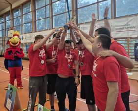Drużyna w czerwonych koszulkach z medalami na szyjach, wznosząca wspólnie duży złoty puchar w hali sportowej. Wszyscy są uśmiechnięci i radośnie celebrują. Po lewej stronie stoi maskotka w przebraniu czerwono-niebieskiego ptaka. W tle widać siatkę okienną i banery.