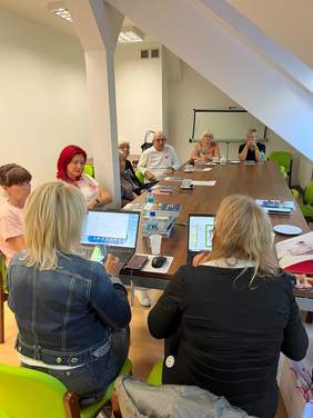 Pokaż zdjęcie: Przy stole siedzą uczestnicy spotkania oraz przy komputerach pracownicy CIDON