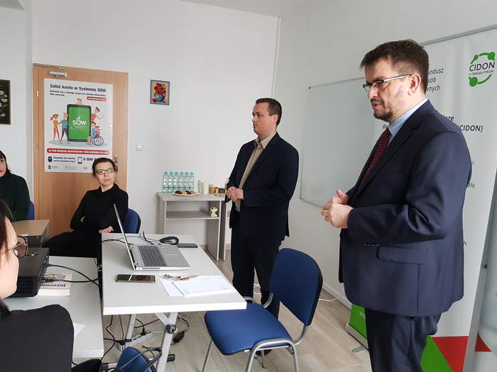 Pokaż zdjęcie: Dyrektor Oddziału stoi w sali konferecyjnej i wita gości, obok stoi pracownik Oddziału, jest laptop, biurko
