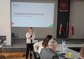 Pokaż zdjęcie: Sala konferencyjna, na ścianie wyświetlana jest prezentacja z dostępności architektonicznej, prelegentem jest kobieta ubrana w białą bluzkę i czarne spodnie