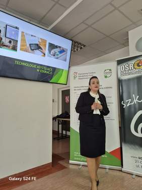 Pokaż zdjęcie: Kobieta stoi przy rollupie z logo centrum informacyjno-doradczego CIDON, w tle slajd z prezentacją.