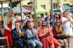Pokaż zdjęcie: Pod namiotem na krzesłach siedzą elegancko ubrane osoby, które klaszczą