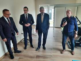 Pokaż zdjęcie: W sali do rehabilitacji znajduje się czterech mężczyzn. Trzech panów jest ubranych w garnitury. Oglądają pomieszczenie. Obok nich stoi fotograf z aparatem w dłoniach. 