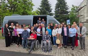 Pokaż zdjęcie: Duża grupa osób stoi na tle busa. Pośród tych osób osoby na wózkach inwalidzkich.
