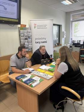 Pokaż zdjęcie: Dwóch pracowników PFRON prowadzi rozmowę informacyjną ze starszymi mieszkańcami przy biurku w pomieszczeniu urzędowym. Na stole widoczne są broszury i materiały dotyczące wsparcia osób z niepełnosprawnościami