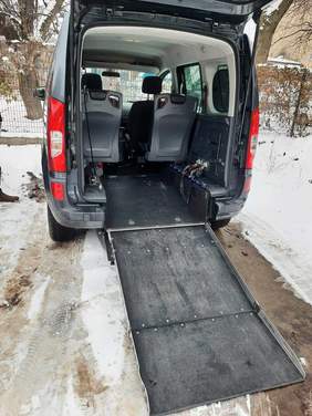 Pokaż zdjęcie: Na parkingu zaparkowany jest samochód dostosowany do przewozu osoby niepełnosprawnej poruszającej się na wózku inwalidzkim. Tylna klapa pojazdu jest otwarta i widać jego wnętrze. Obok auta leży topniejący śnieg. 