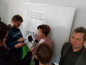 Pokaż zdjęcie: Elegancko ubrana kobieta, przed nią mikrofony. W tle baner z logo PFRON oraz z napisem wszyscy jesteśmy tacy sami. 
