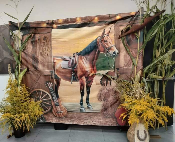 Pokaż zdjęcie: Scenografia w stylu westernowo-farmerskim na jasnej podłodze. W centrum znajduje się obraz z brązowym koniem na tle zachodu słońca, wkomponowany w ścianę przypominającą stodołę z ciepłymi światełkami u góry. Po bokach stoją jesienne dekoracje: łodygi kukurydzy, żółte kwiaty, koło wozu, gitara, dynia oraz worek z podkową.