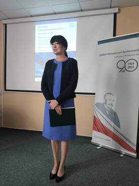 Pokaż zdjęcie: Kobieta w niebieskiej sukience i czarnej marynarce otwiera konferencję.