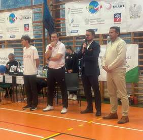 Czterech dorosłych mężczyzn stoi w rzędzie na hali sportowej przed stołem sędziowskim i banerami. Wszyscy są ubrani w stroje codzienne lub sportowe, z wyjątkiem jednego mężczyzny w garniturze. Prawdopodobnie są to organizatorzy lub sędziowie zawodów.