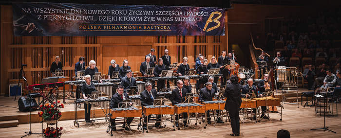 Pokaż zdjęcie: Scena koncertowa z dużym zespołem perkusyjnym Polskiej Filharmonii Bałtyckiej. Muzycy stoją przy marimbach, ksylofonach i wibrafonach, dyrygent prowadzi wykonanie. Nad sceną baner noworoczny z życzeniami i logo PFB.