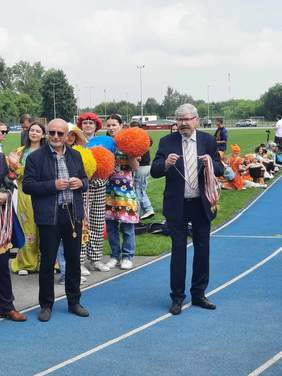 Pokaż zdjęcie: Na boisku sportowym stoją różne osoby. Na środku jest pan, który trzyma w dłoni wiele medali. 