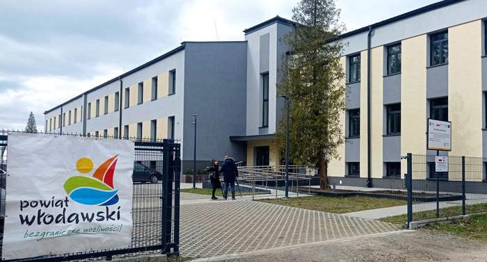 Pokaż zdjęcie: Zdjęcie dwupiętrowego budynku w kolorze szaro-beżowym, z dużą ilością okien. W budynku mieściła się wcześniej szkoła.