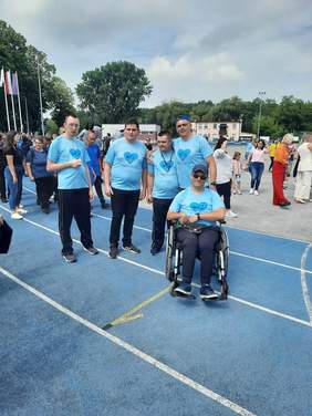 Pokaż zdjęcie: Na boisku sportowym jest pięciu panów: czterech stoi, a jeden siedzi na wózku inwalidzkim. Są ubrani w takie same błękitne koszulki. 