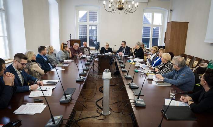 Pokaż zdjęcie: Przy okrągłym stole, w sali ratusza, znajdują się osoby biorące udział w spotkaniu
