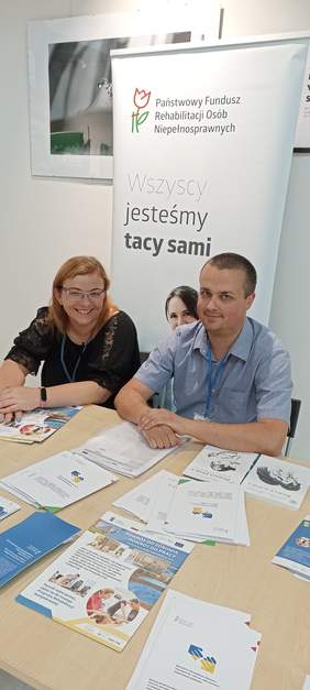 Pokaż zdjęcie: Przedstawiciele Oddziału Lubuskiego PFRON na tle baneru PFRON siedza przy ławce, na której są rozłożone materiały informacyjne