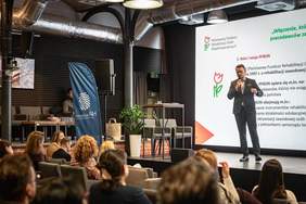 Przedstawiciel PFRON omawia instrumenty wsparcia pracodawców zatrudniających osoby z niepełnosprawnościami podczas konferencji „Biznes i Inkluzja”. Wystąpienie odbywa się na scenie przed zgromadzoną publicznością, a na ekranie widoczna jest prezentacja dotycząca roli i misji PFRON.