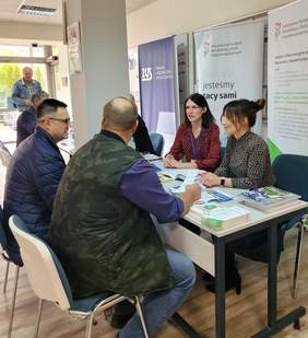 Pokaż zdjęcie: dwóch mężczyzn siedzi tyłem przed nimi za biurkiem dwie kobiety udzielają porad.