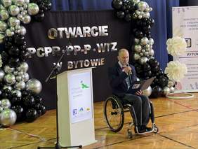 Pokaż zdjęcie: Otwarta sala z czarno-srebrnymi balonami, napis „Otwarcie PCPR · PP-P · WTZ w Bytowie”, mównica z logo Powiatu Bytowskiego, obok mężczyzna na wózku w garniturze mówi do mikrofonu i trzyma teczkę WTZ.
