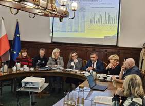 Pokaż zdjęcie: Grupa osób siedzi przy owalnym stole w sali konferencyjnej. Na ścianie wyświetlana jest prezentacja z wykresami i tabelami. W tle widoczne są flagi Polski i Unii Europejskiej. Spotkanie ma oficjalny, roboczy charakter.