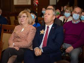 Pokaż zdjęcie: Widownia, osoby w różnym wieku siedzą na krzesłach i patrzą w kierunku sceny, na pierwszym planie dwie osoby - elegancko ubrana kobieta i mężczyzna.