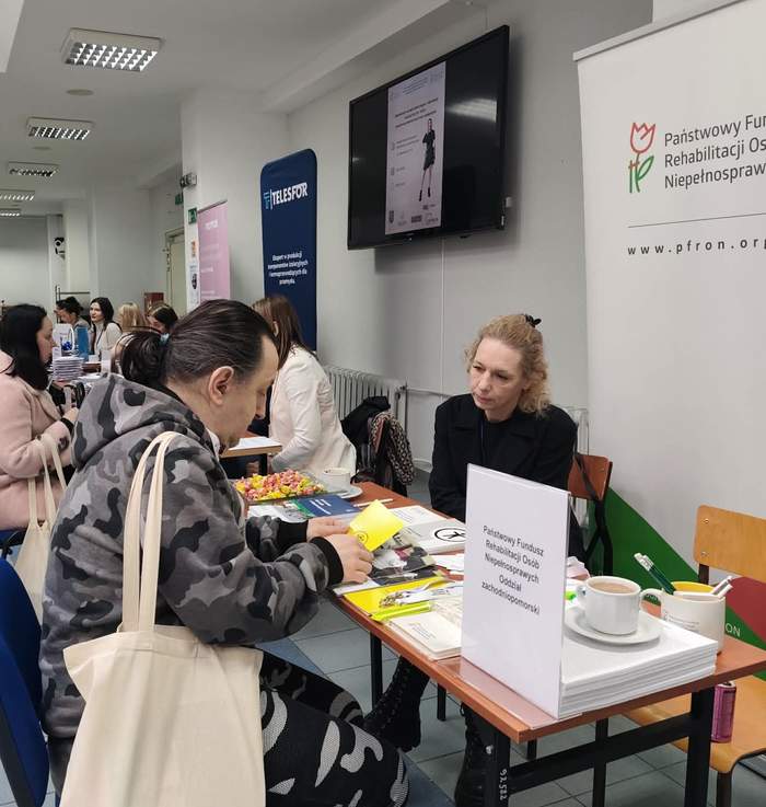 Pokaż zdjęcie: Zdjęcie przedstawia stoisko PFRON w trakcie trwania wydarzenia. Przy stoliku siedzi kobieta (przedstawicielka instytucji) w czarnym ubraniu, która rozmawia z mężczyzną siedzącym naprzeciwko niej. Mężczyzna w bluzie moro przegląda żółtą ulotkę. Na stoliku, obok materiałów promocyjnych i słodyczy, stoi biała filiżanka z kawą. W tle widać długi korytarz z innymi stoiskami targowymi (m.in. firmy Telesfor), przy których siedzą ludzie. Na ścianie powyżej stoiska zawieszony jest ekran telewizyjny wyświetlający plakat informacyjny.