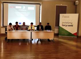 Pokaż zdjęcie: Organizatorzy konferencji.  Siedzą za stołem na tle ekranu z tytułem konferencji. 