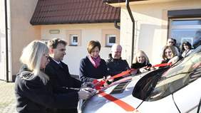 Pokaż zdjęcie: Grupa ośmiu osób stoi przed budynkiem, obok którego znajduje się srebrny autobus z czerwoną kokardą na masce pojazdu. Kilka osób, które otaczają pojazd, przecina z różnych stron kokardę nożyczkami.