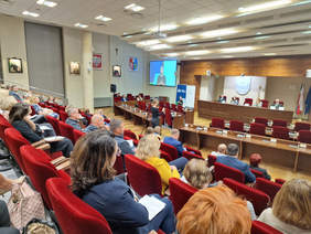 Widok na sale konferencyjną z boku i z góry, widać uczestników, za stołem prezydialnym na przodzie siedzą 4 osoby