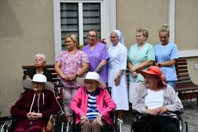 Pokaż zdjęcie: Przed budynkiem znajduje się dziewięć kobiet, w tym siostra zakonna oraz trzy kobiety poruszające się na wózku i mające założony na głowie kapelusz. Wzdłuż budynku ustawiona jest drewniana ławka.