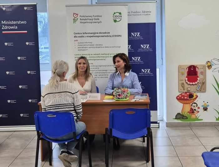 Pokaż zdjęcie: Przy biurku informacji kobiecie udzielają pracownice funduszu. Na biurku leżą ulotki, katalogi, foldery.