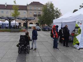 Pokaż zdjęcie: Na dziedzińcu ustawione są namioty informacyjne, przed którymi stoją ludzie. W tle widać drzewa i fontanny. 