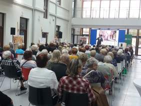 Pokaż zdjęcie: Sala konferencyjna, osoby siedzące tyłem w oddali scena, na niej stoi osoba z mikrofonem. 