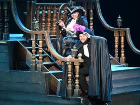 Pokaż zdjęcie: Dwóch aktorów w teatralnych kostiumach piratów na drewnianej scenografii statku. Jeden trzyma się barierki, drugi siedzi za sterem w kapeluszu z piórem.