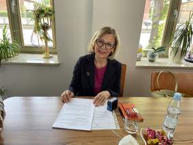 Pokaż zdjęcie: Uśmiechnięta kobieta w blond włosach i okularach, ubrana w ciemną marynarkę siedzi za biurkiem. W ręku trzyma umowę, obok leży pieczątka. Za nią okna, na parapetach stoją kwiaty.