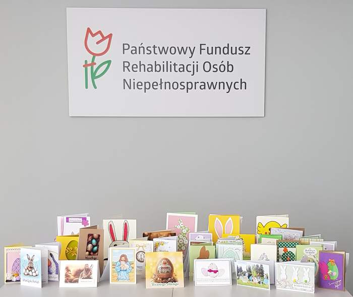 Pokaż zdjęcie: Na jasnoszarym tle u góry znajduje się logo z czerwonym tulipanem i zieloną łodygą oraz napis: Państwowy Fundusz Rehabilitacji Osób Niepełnosprawnych Na dole grafiki stoi rząd rozłożonych, kolorowych kartek wielkanocnych. 