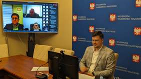 Pokaż zdjęcie: Na zdjęciu widzimy mężczyznę, który siedzi przed laptopem i rozmawia. Z tyłu znajduje się telewizor z wyświetloną konferencją.