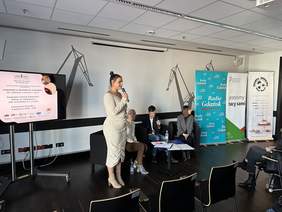 Pokaż zdjęcie: Kobieta w jasnej sukni spiętych włosach mówi do mikrofonu. Po jej lewej duży monitor z logo Szansa Start i tytułem konferencji. Po prawej stronie siedzą trzy osoby na fotelach. W tle rysunki dźwigów stoczni gdańskiej.