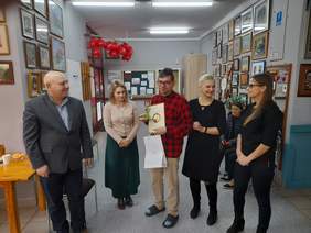 Pokaż zdjęcie: W pracowni artystycznej Warsztatu Terapii Zajęciowej pozuje do pamiątkowego zdjęcia 5 osób. Pośrodku stoi mężczyzna w czerwonej koszuli w kratę, w ręku trzyma dyplom oraz siatkę z nagrodami. Po lewej stronie zdjęcia stoją pracownicy PFRON mężczyzna i kobieta, po prawej dwie kobiety pracujące w WTZ w Chełmie.