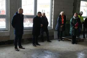 Pokaż zdjęcie: Mężczyzna w płaszczu i garniturze mówi do kobiety. Obok stoją dwaj mężczyźni również w płaszczach. Wszyscy znajdują się na piętrze budynku w stanie surowym.