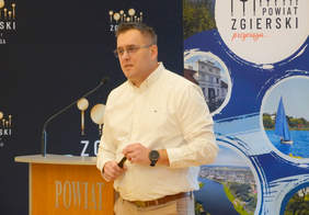 Pokaż zdjęcie: Mężczyzna prowadzi prelekcję. Za nim widać mównicę z mikrofonem i banery informacyjne. Jesy ubrany w białą koszulę. 