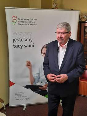 Pokaż zdjęcie: Na pierwszym planie stoi mężczyzna w garniturze. Za nim znajduje się baner Państwowego Funduszu Rehabilitacji Osób Niepełnosprawnych. 