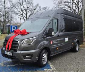 Pokaż zdjęcie: Ciemnoszary bus (duży van) zaparkowany na zewnątrz, z dużą czerwoną kokardą i wstążkami ułożonymi na masce z przodu. Na zdjęciu widać ujęcie z przodu i z boku, z przyciemnianymi oknami w części pasażerskiej. Bus stoi na niebieskim miejscu parkingowym; obok jest znak parkingu z literą „P”, a w tle widać bezlistne drzewa. Na boku pojazdu są logotypy: Miasto i Gmina Grabów nad Prosną oraz Państwowego Funduszu Rehabilitacji Osób Niepełnosprawnych. 