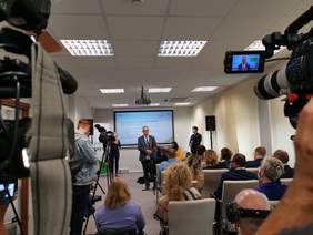 Pokaż zdjęcie: Sala konferencyjna. Nagrywane przez media spotkanie z wygłaszającym wystąpienie mężczyzną w szarym garniturze. Przed nim zgromadzona na krzesłach grupa kilkunastu osób. W tle prezentacja multimedialna.