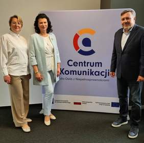 Pokaż zdjęcie: Dwie kobiety i jeden mężczyzna elegancko ubrani stoją przy rollupie z napisem i logo Centrum Komunikacji.