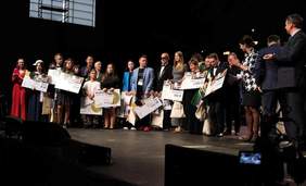 Pokaż zdjęcie: Oświetlona scena. Na scenie stoją uśmiechnięci laureaci oraz jury i organizatorzy. 