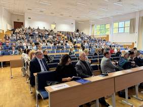 Pokaż zdjęcie: Duża aula z licznymi rzędami miejsc; uczestnicy konferencji siedzą w ławkach, część przegląda dokumenty. Jasne wnętrze z dużymi oknami po prawej stronie, podwyższenie z tyłu sali.