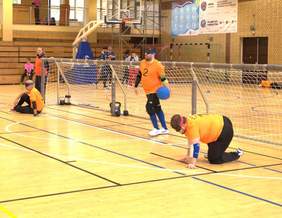 Zawodnicy w pomarańczowych koszulkach rozgrywają mecz goalball na hali sportowej. Jeden z nich trzyma niebieską piłkę, a drugi klęczy przed bramką.